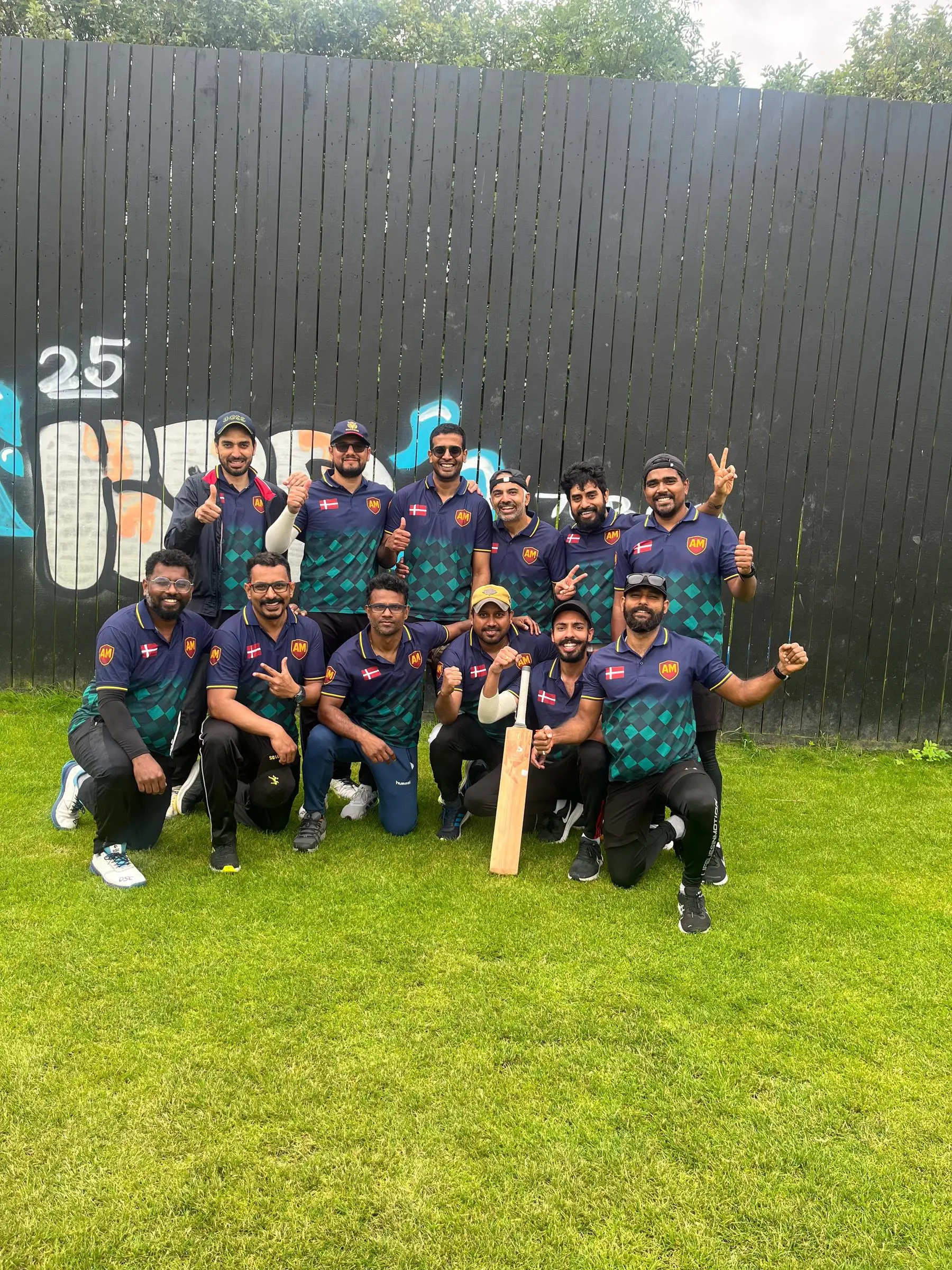 The cricket squad posing with a bat in team kit against a painted fence.