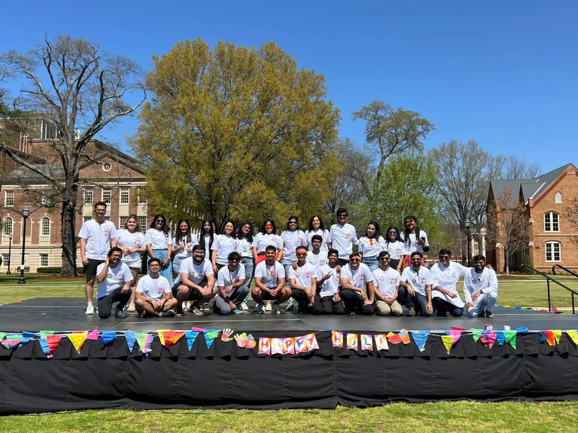 The NSA ISAT team on an outdoor stage in the white event t-shirt, a Happy Holi banner along the front.