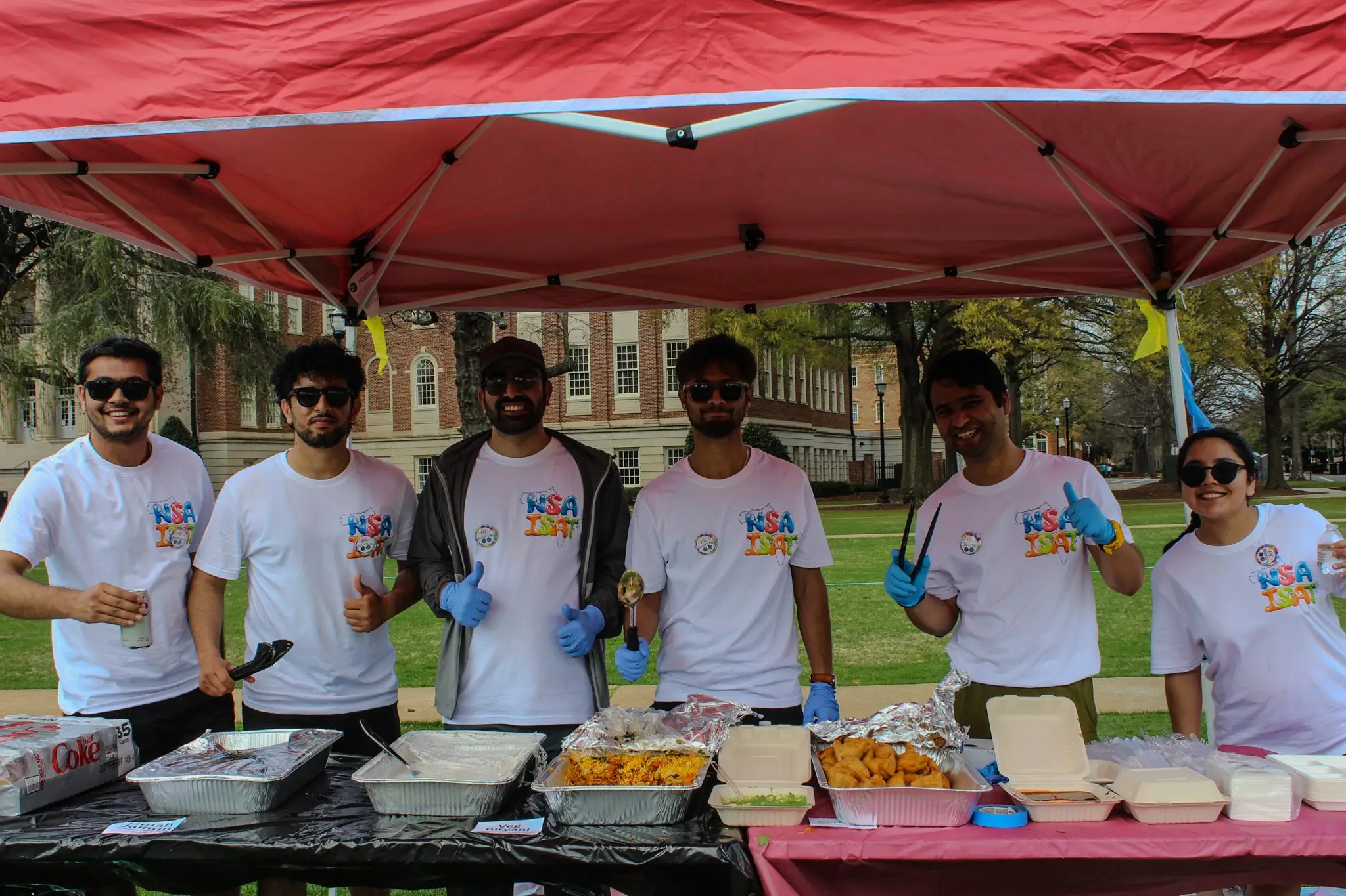 Volunteers in NSA ISAT event t-shirts serving food at a stall under a red canopy.