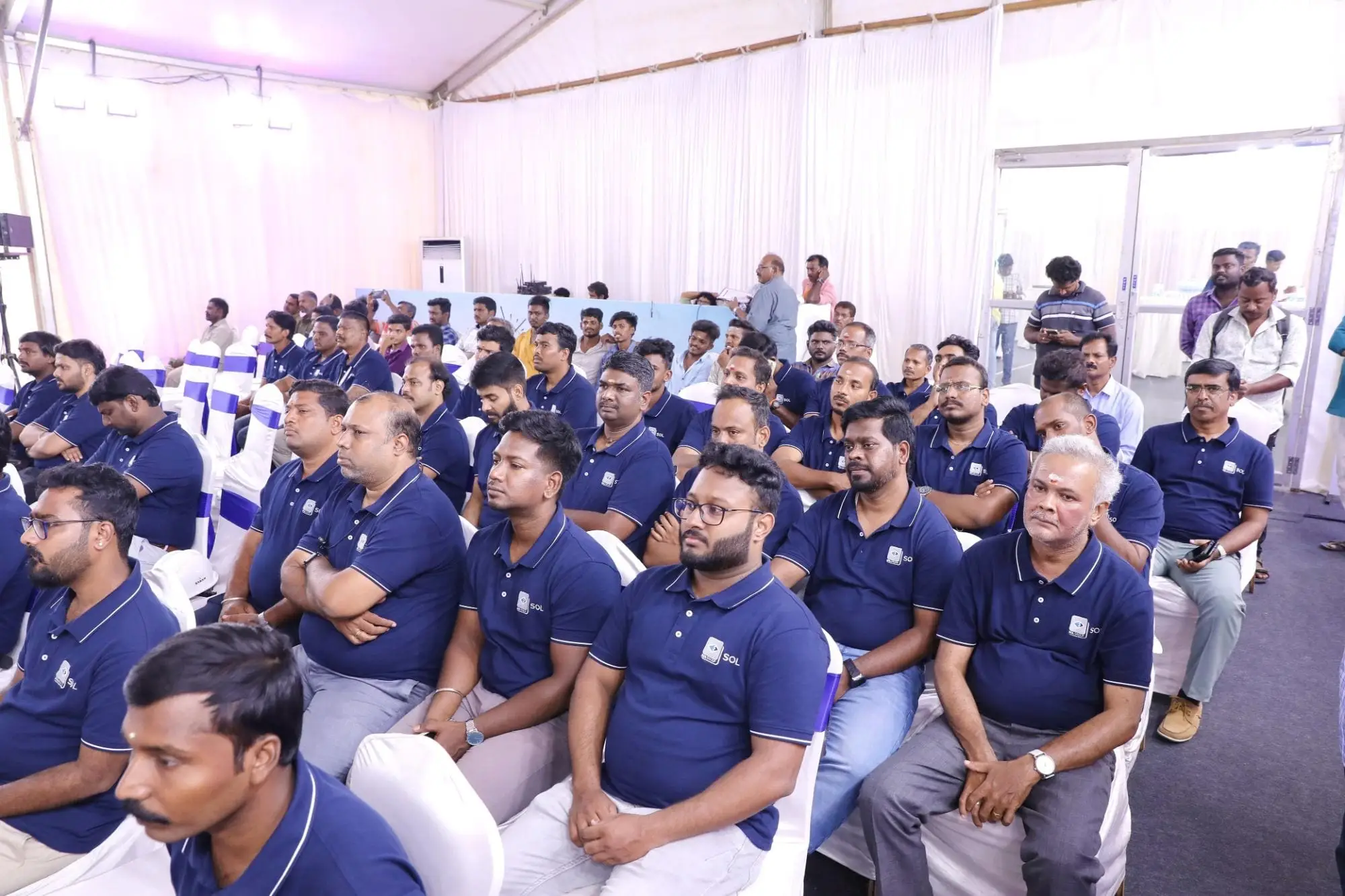 The ceremony audience under the marquee, row after row in the navy SOL polo.