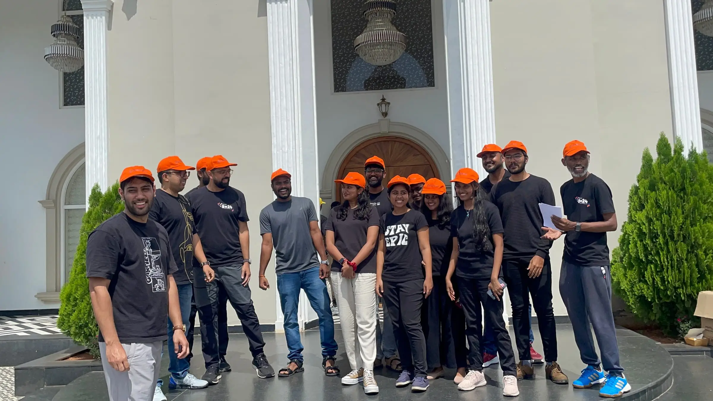The same team in black tees and orange caps, gathered in front of the building's arched doorway.