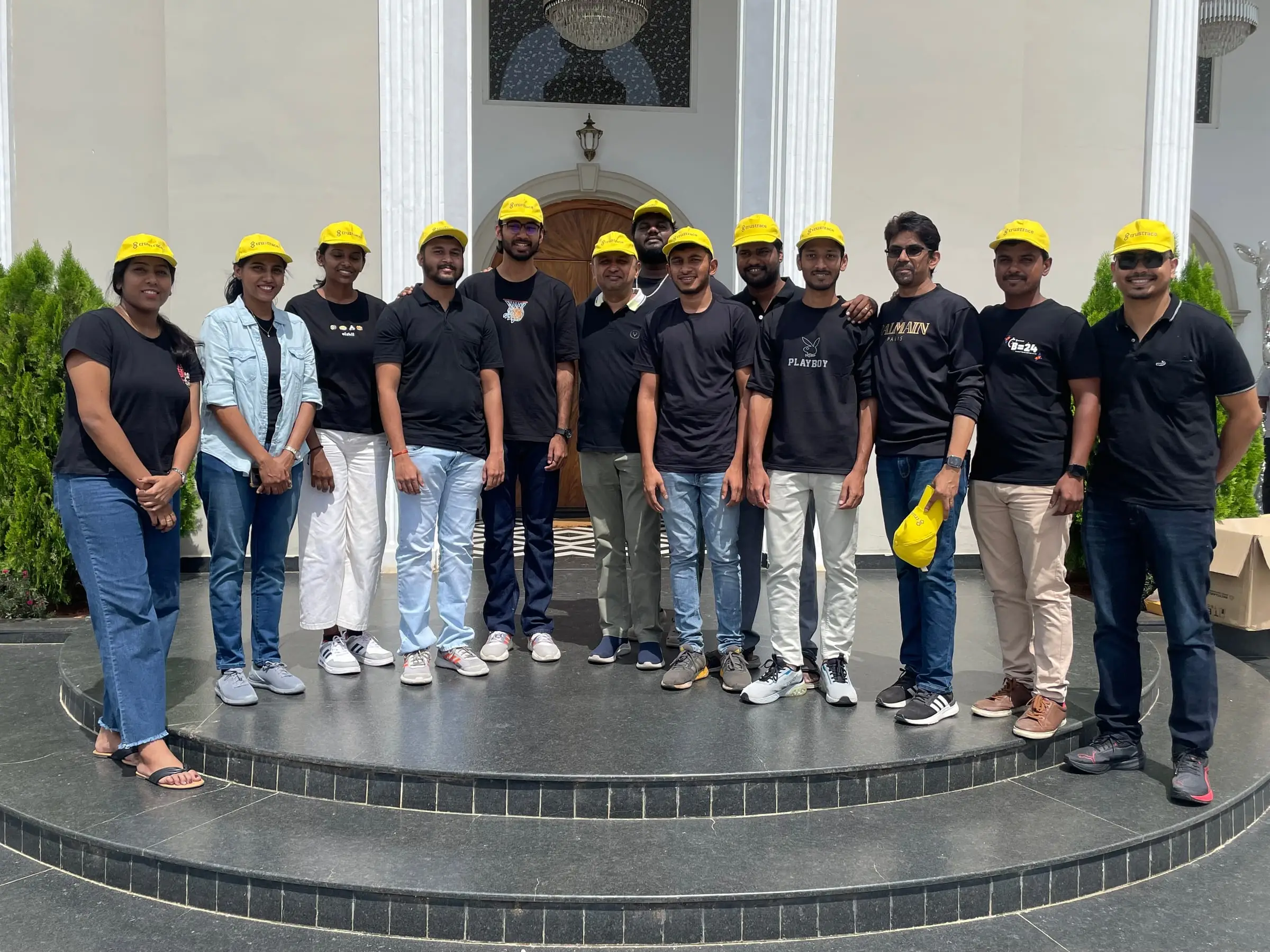 A team lined up on the steps of a white building in black printed tees and yellow caps.