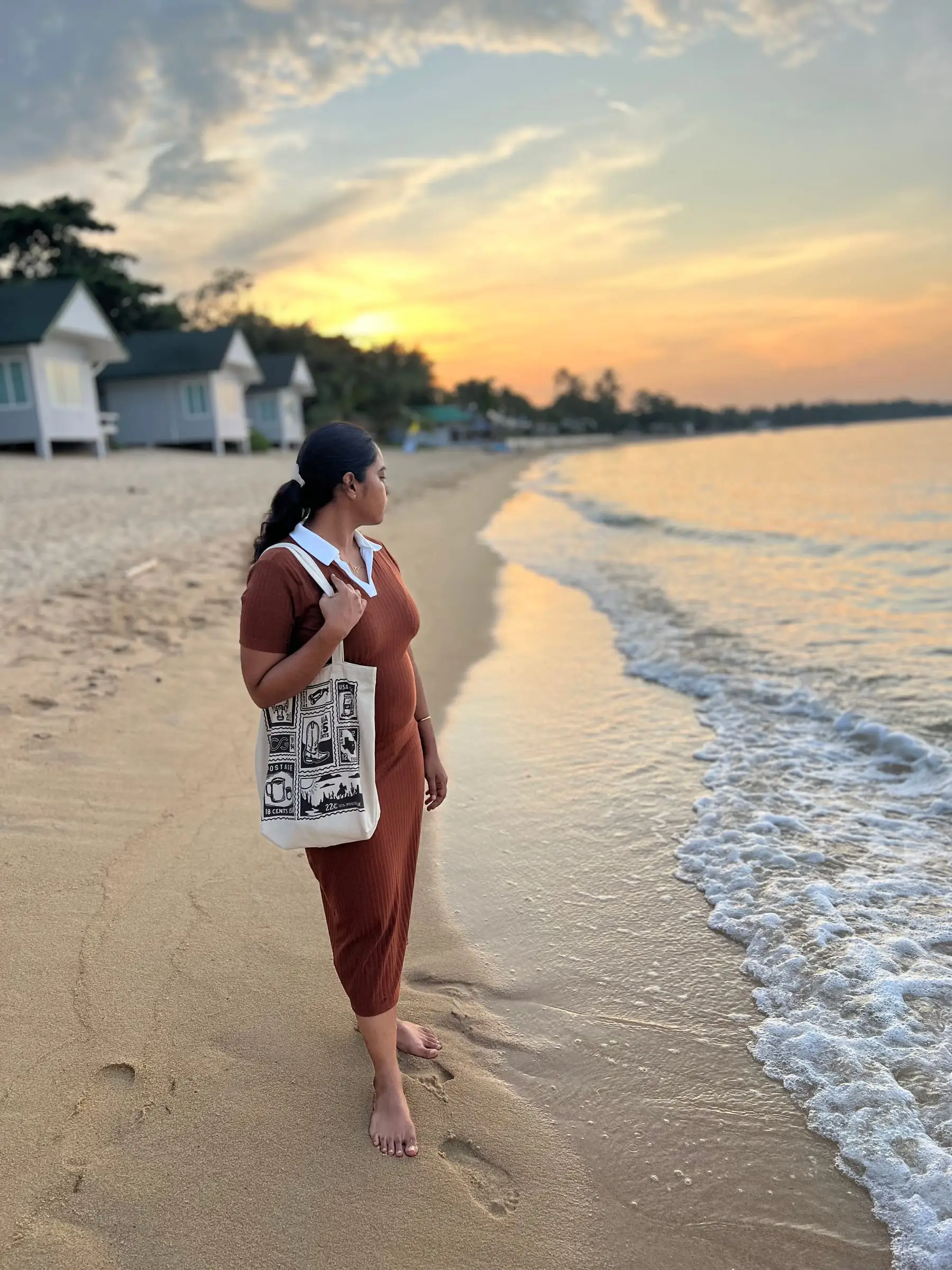 A woman walking along the shore at sunset with a canvas tote on her shoulder, a postage-stamp print across the bag.