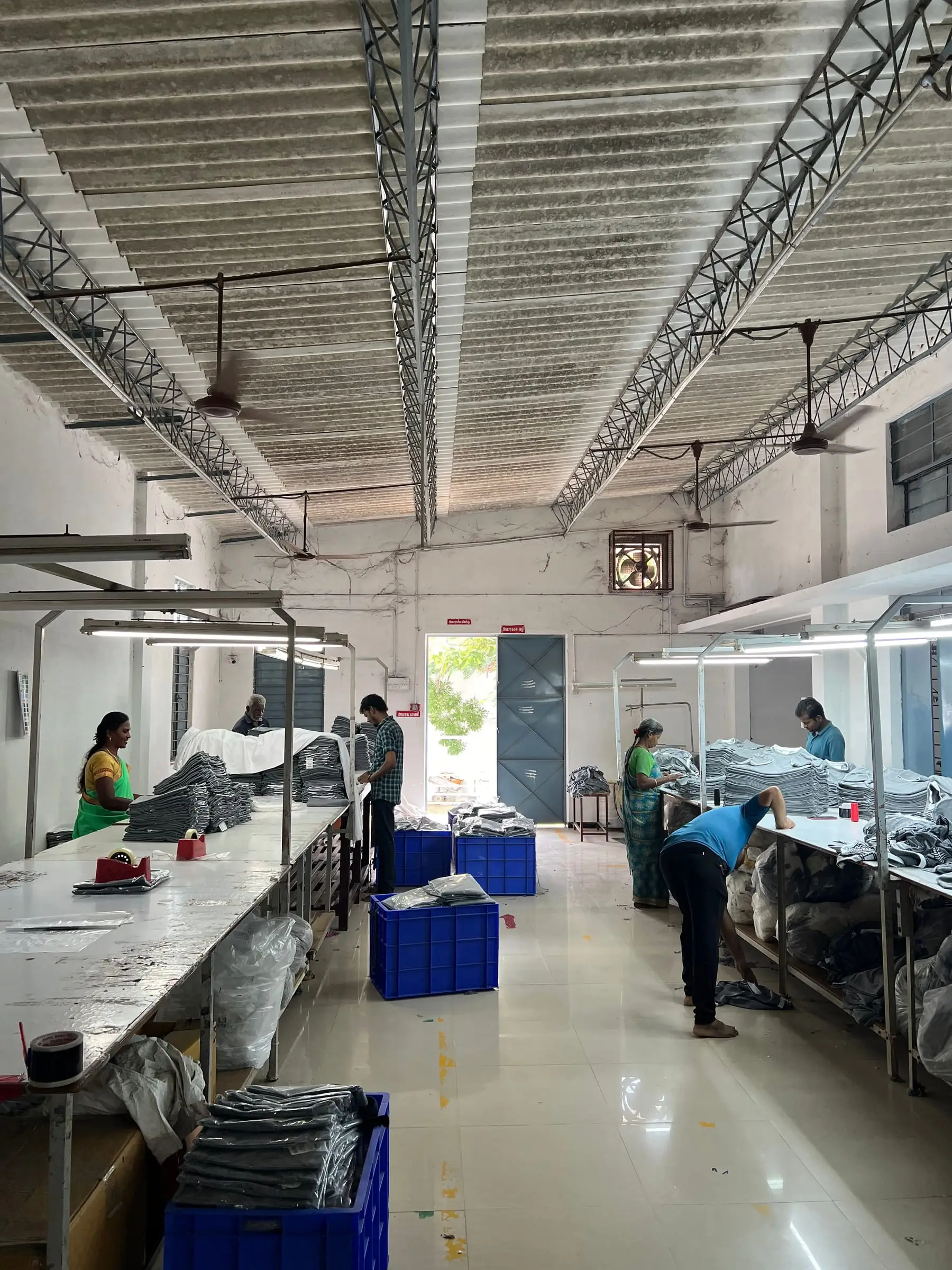 The finishing floor at Tiruppur seen end to end — cutting and folding tables, crates and stacked stock under the roof trusses.