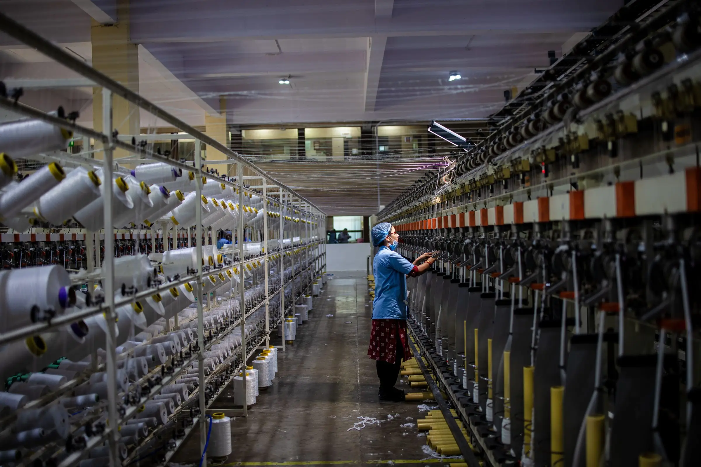 A long aisle of yarn-winding frames, white cotton cones racked on the left and spindles running down the right, with an operator checking a feed mid-aisle.