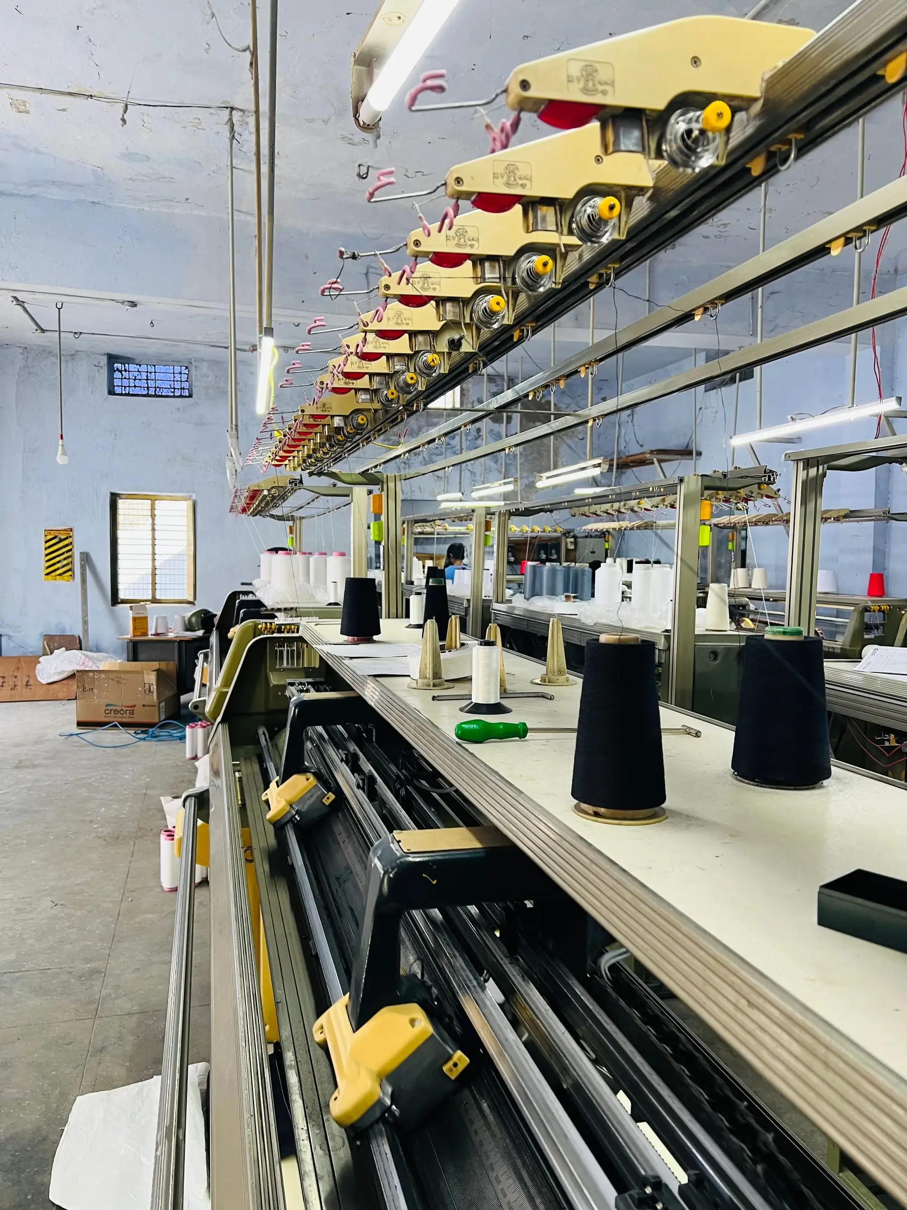 A row of flat-knitting machines running the length of the knitting hall, yarn cones lined up along the bed.