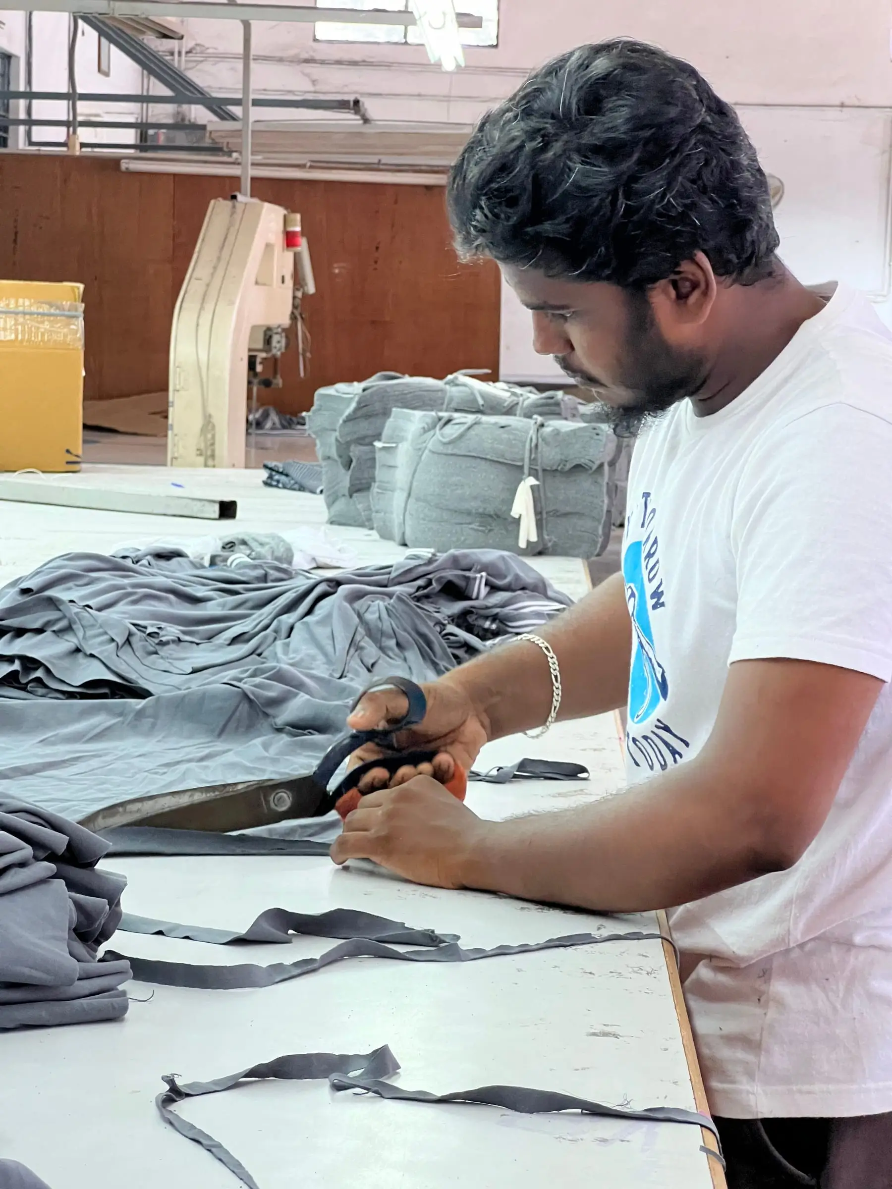 A machinist trimming and squaring grey t-shirt panels with shears at the cutting table.